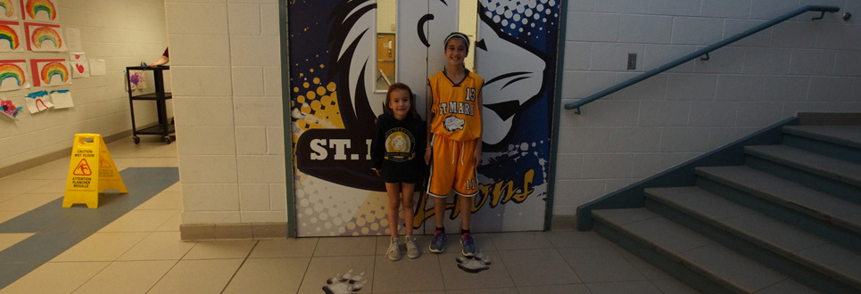 Two female students wearing spirit wear standing in front of gym doors